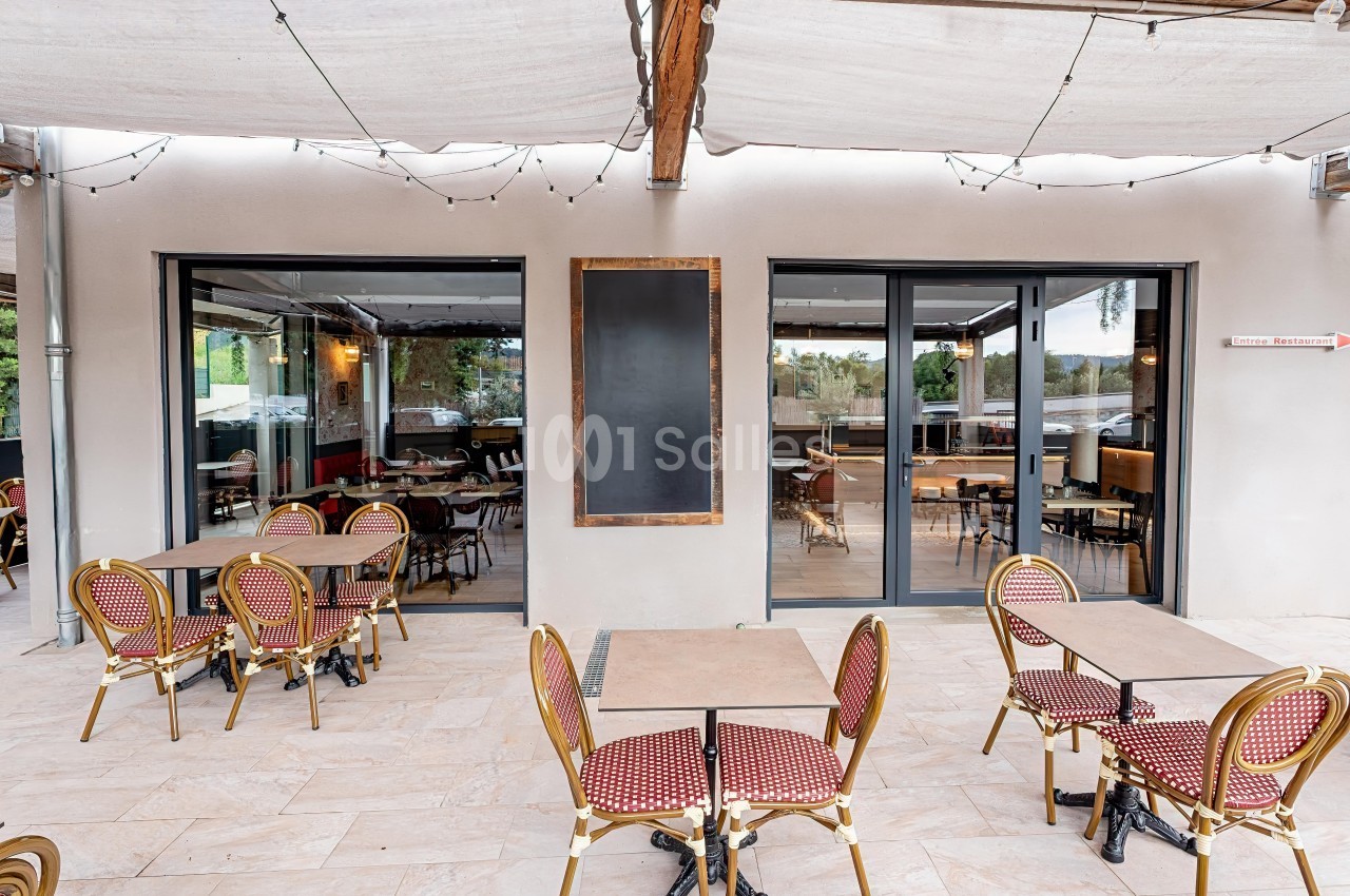 Terrasse de café avec tables et chaises en rotin rouge, devant une façade vitrée et un tableau noir vide.