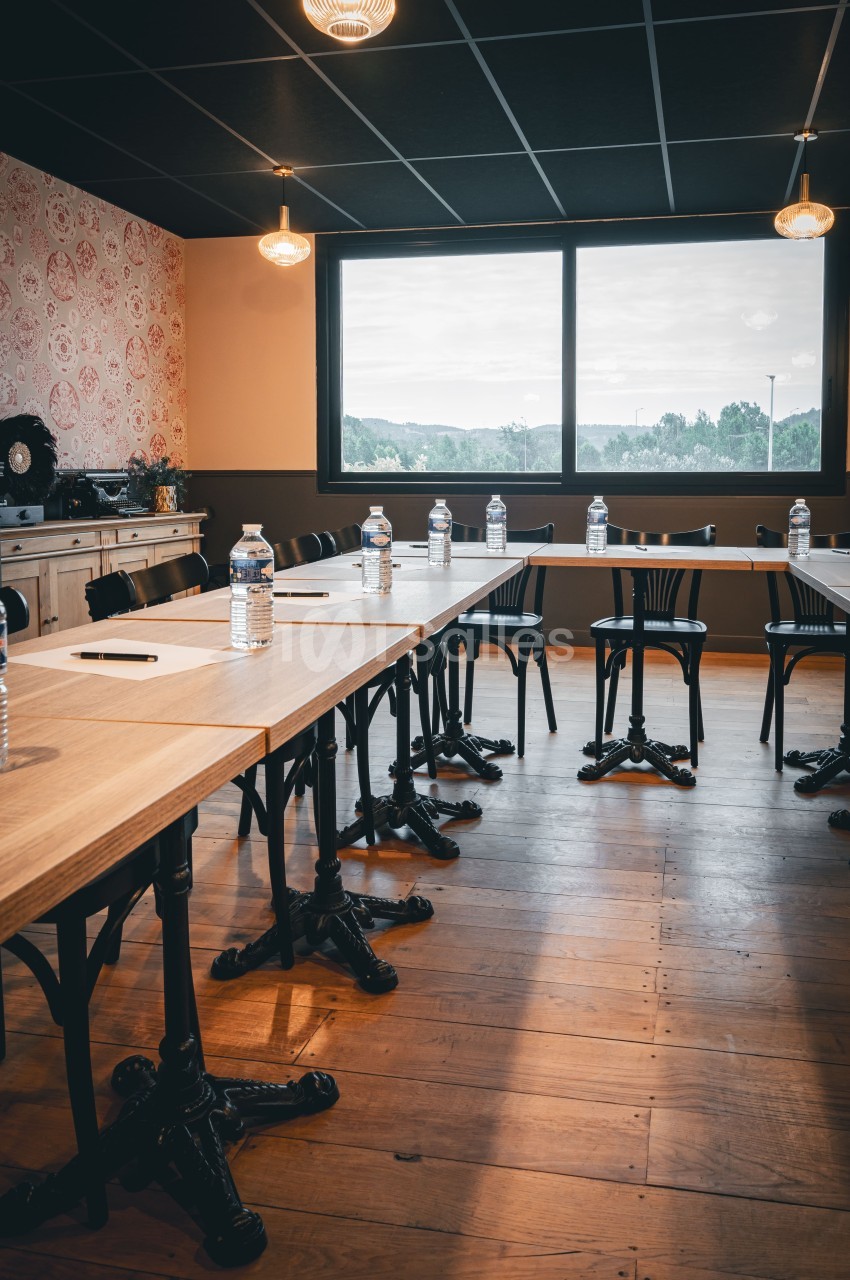 Salle de réunion avec tables en bois, chaises noires, bouteilles d'eau et carnets, éclairée par une grande fenêtre.