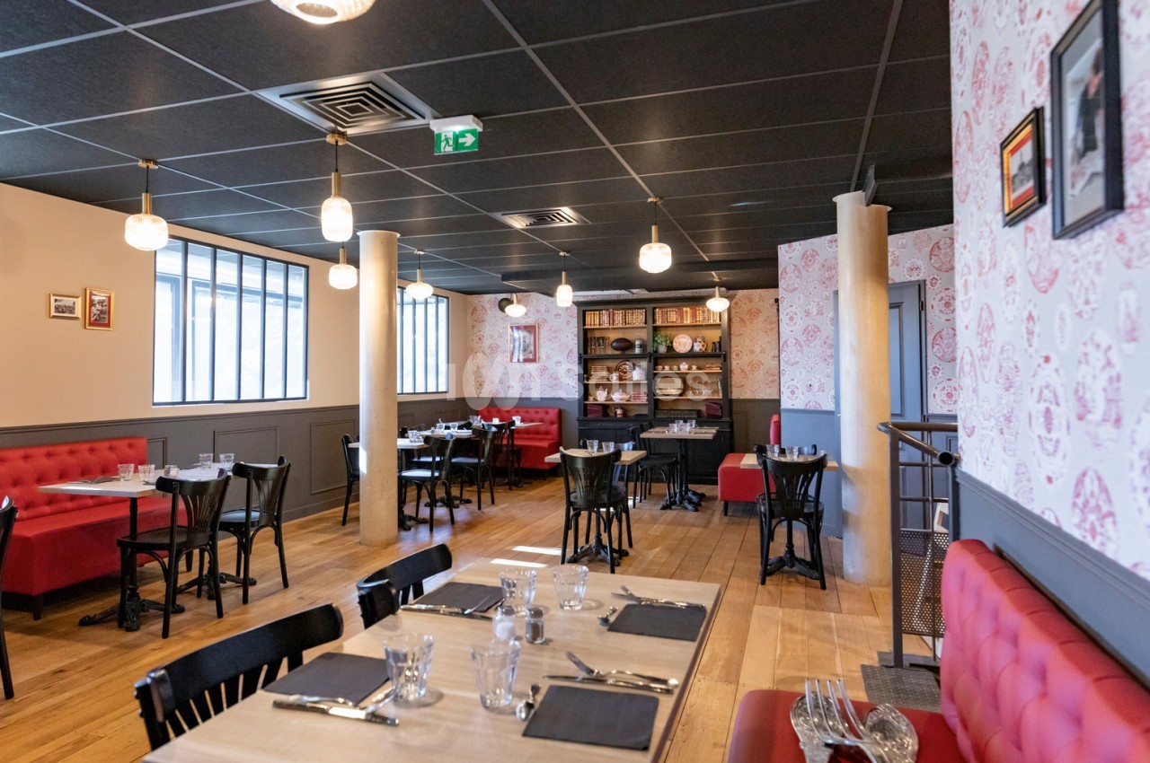 Salle de restaurant lumineuse avec tables dressées, banquettes rouges, étagères décoratives et parquet en bois clair.