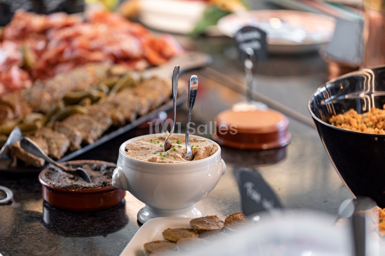 Bol de rillettes avec cuillères, entouré de charcuterie, cornichons et autres plats sur une table de buffet.