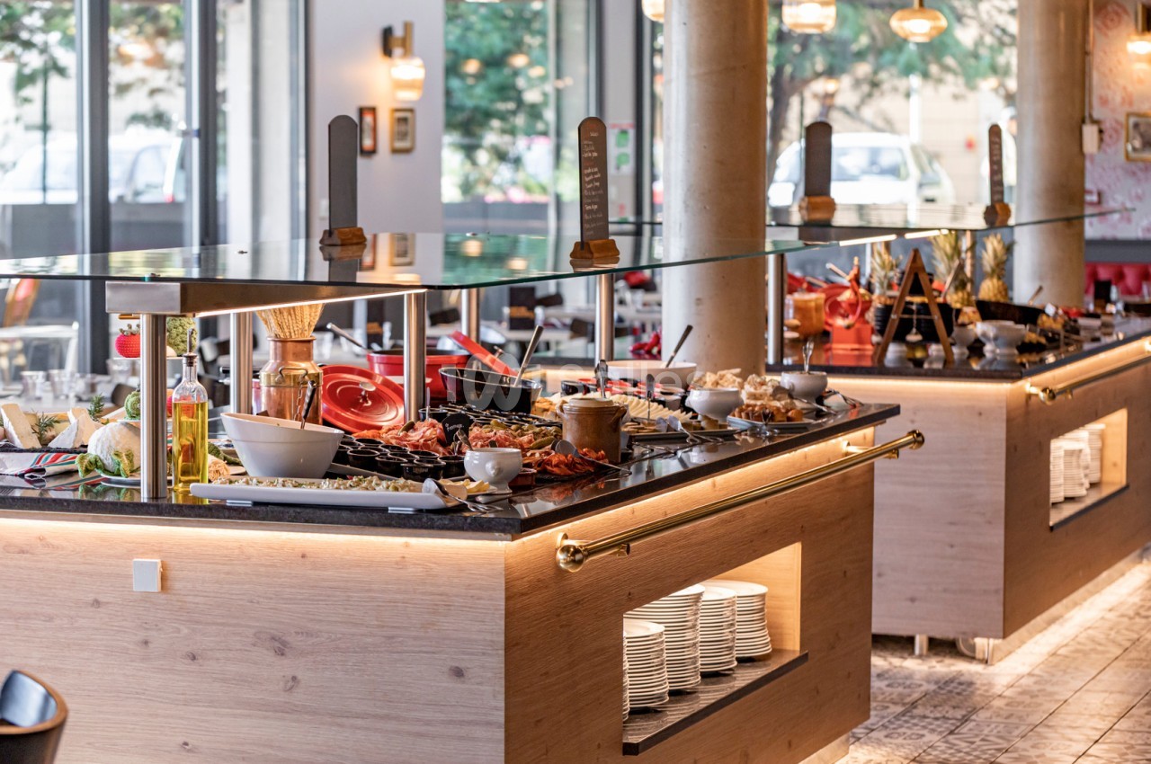 Buffet varié avec plats, condiments et ustensiles disposés sur des comptoirs en bois dans un restaurant lumineux.