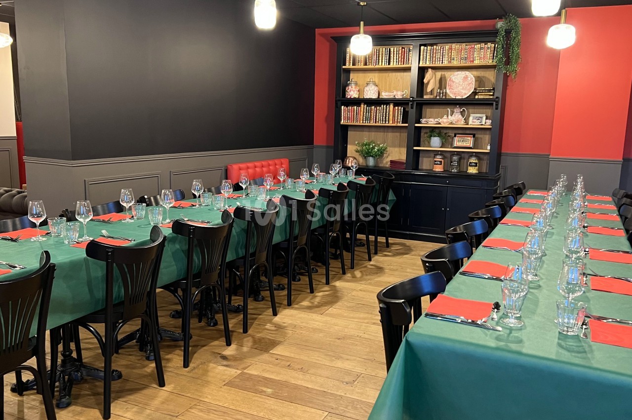 Salle de restaurant aménagée avec deux longues tables dressées, nappes vertes, serviettes rouges et étagères en arrière-plan.