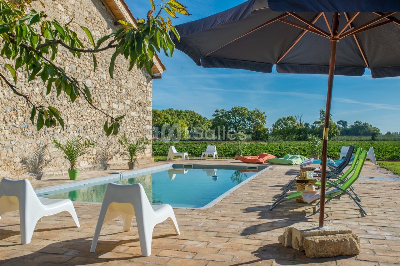 Piscine extérieure entourée de chaises longues et parasols, située près d'une maison en pierre et d'un paysage verdoyant.