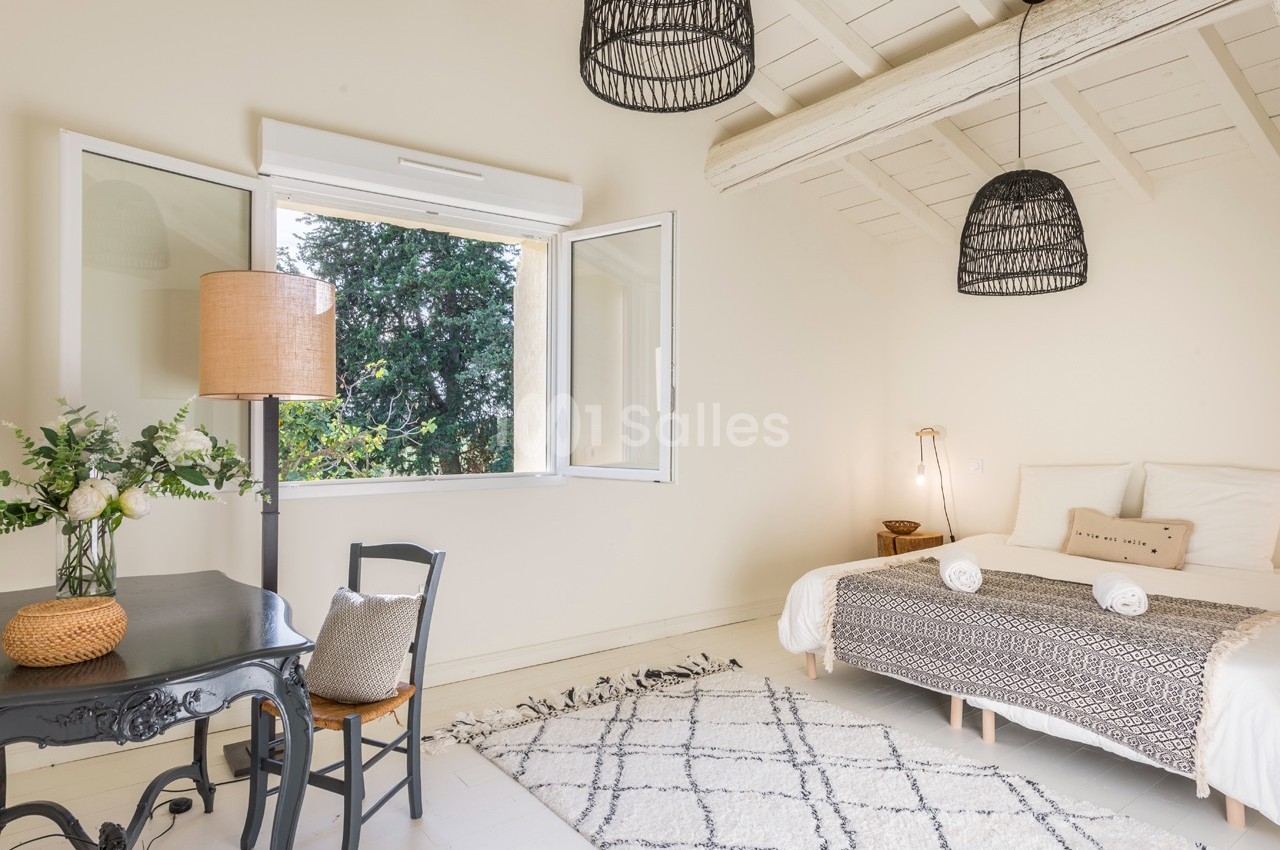 Chambre lumineuse avec lit double, bureau noir, lampe en osier, fenêtre ouverte et plafond avec poutres apparentes.