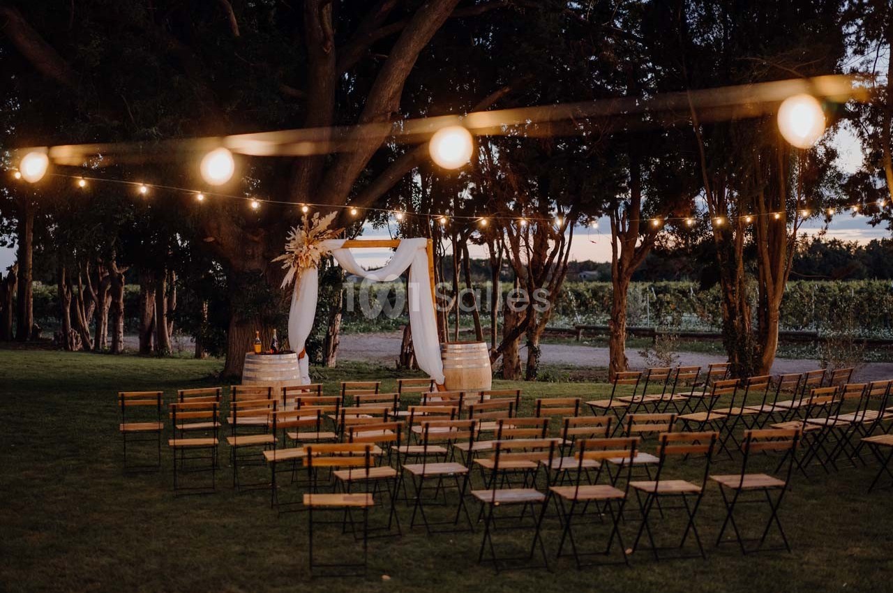 Chaises alignées devant une arche décorée de tissu blanc et de fleurs, éclairée par des guirlandes lumineuses dans un jardin…