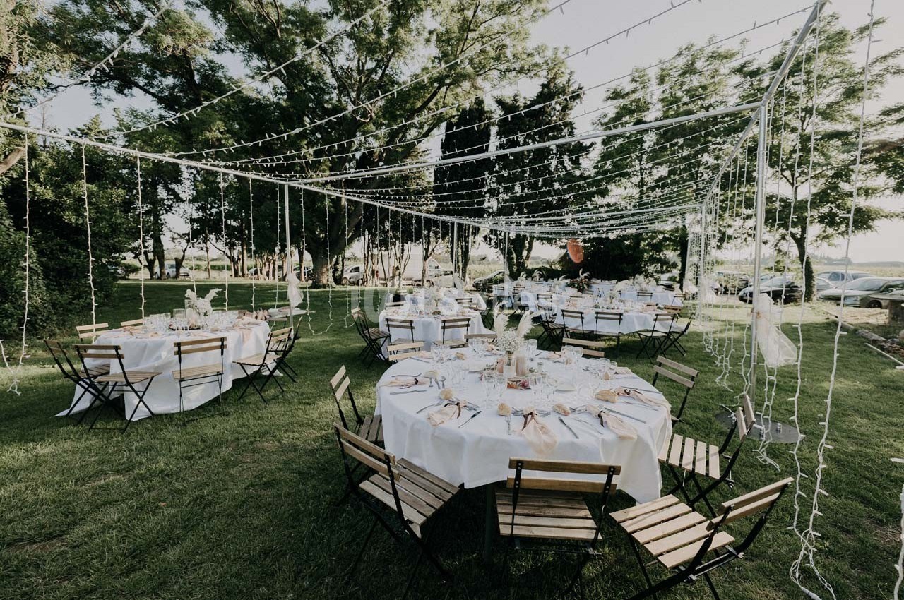 Tables rondes dressées avec nappes blanches et chaises pliantes en bois, disposées sous des guirlandes lumineuses dans un…