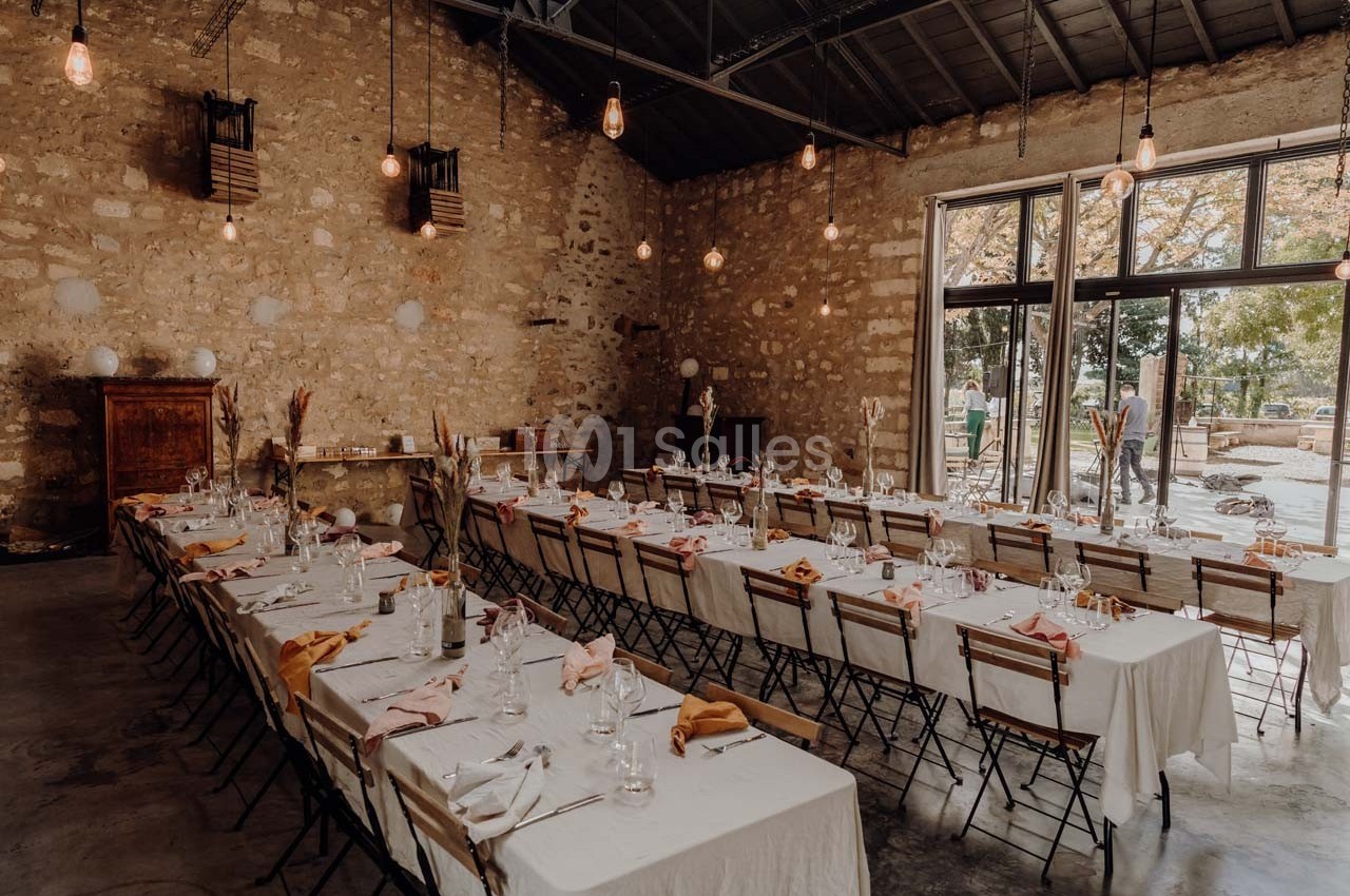 Salle en pierre avec de grandes tables dressées pour un repas, éclairée par des ampoules suspendues et une lumière naturelle.
