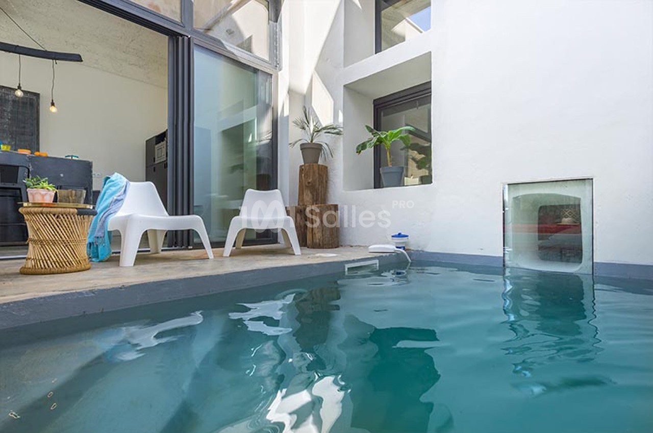 Piscine intérieure moderne avec chaises blanches, table en rotin et plantes près de grandes baies vitrées.