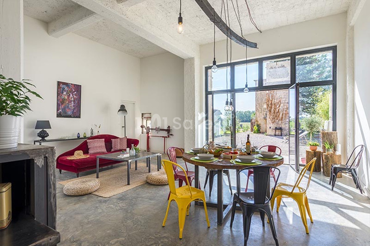 Espace de vie lumineux avec table dressée, canapé rouge, chaises colorées et grandes baies vitrées donnant sur un jardin.