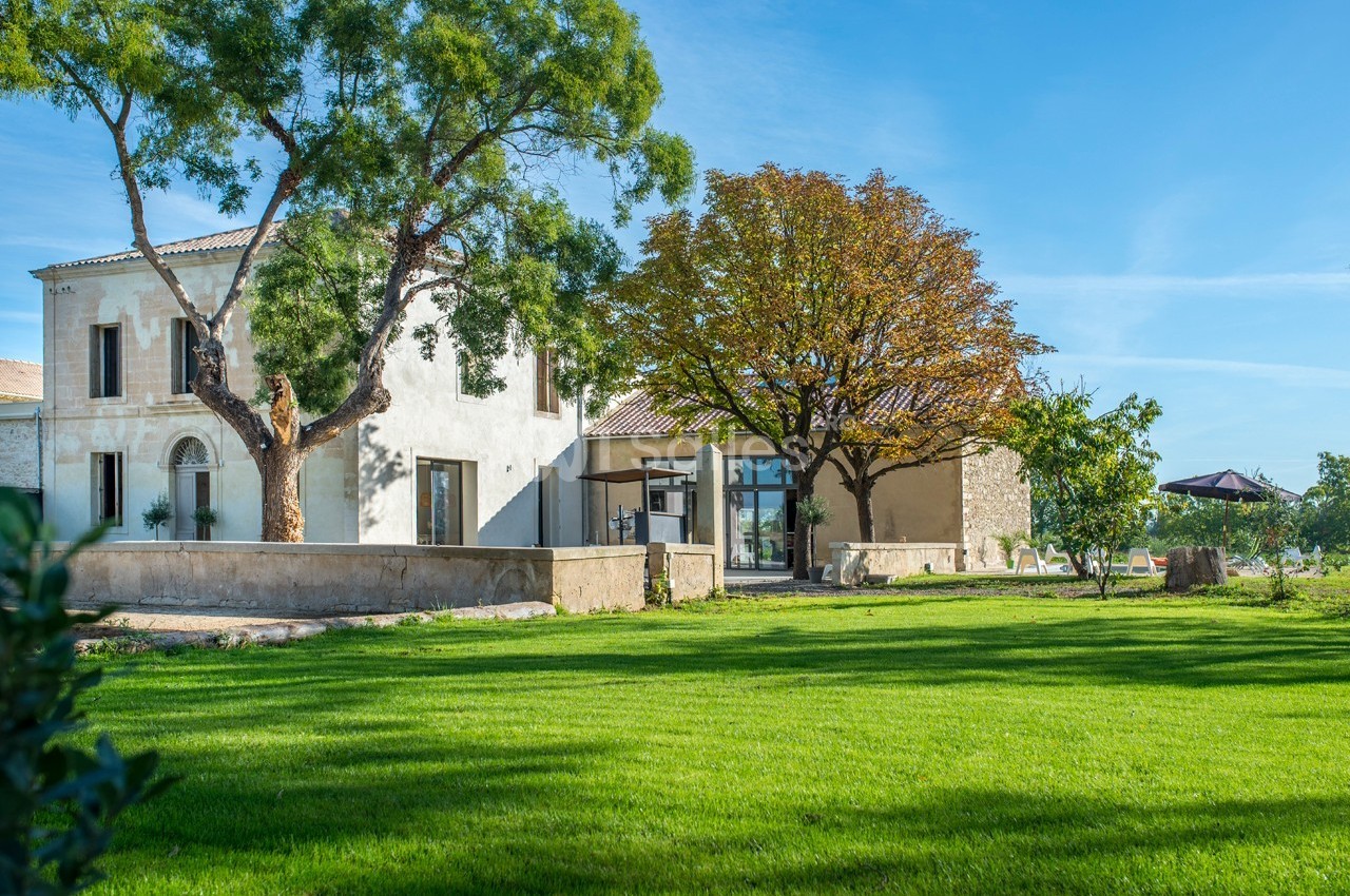 Maison en pierre entourée d'un jardin verdoyant, avec des arbres et un ciel bleu en arrière-plan.
