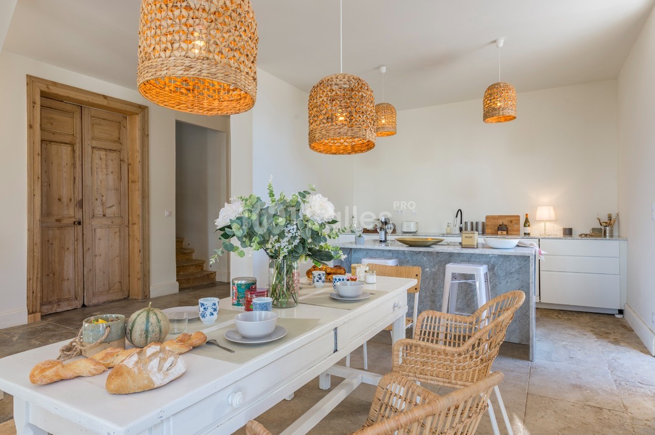 Salle à manger lumineuse avec table dressée, chaises en rotin, suspensions en osier et cuisine ouverte en arrière-plan.