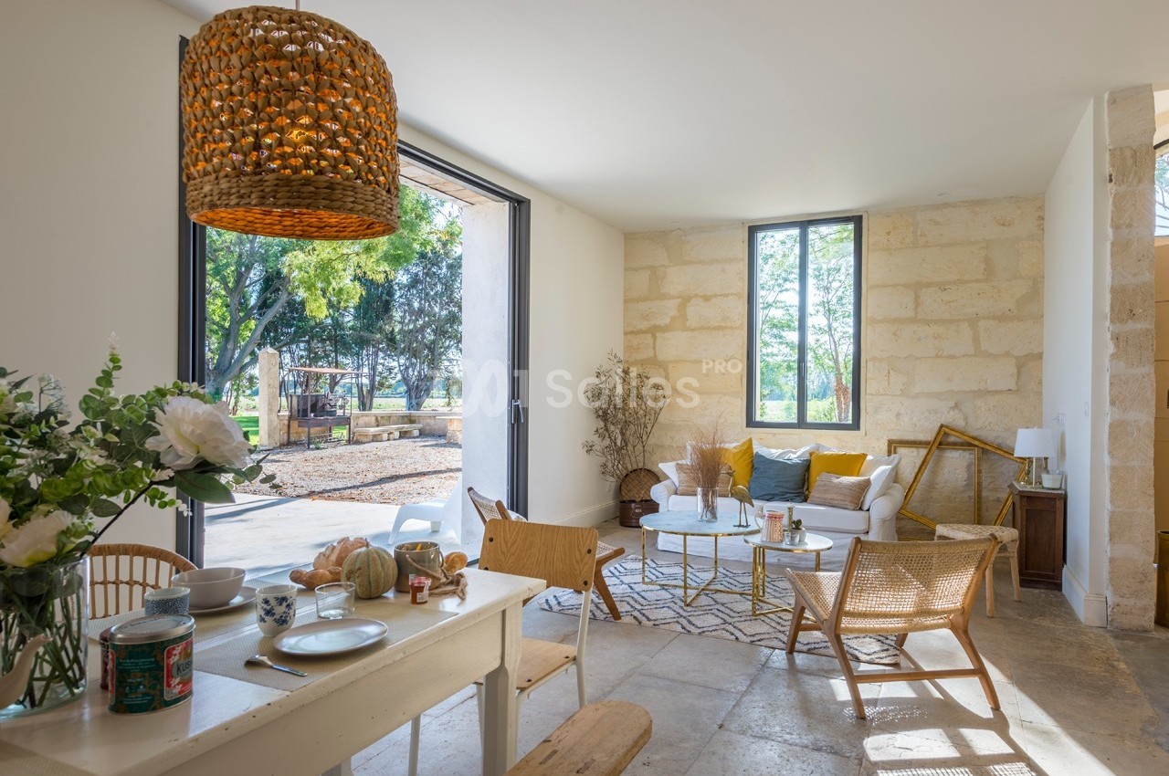 Salon lumineux avec murs en pierre, mobilier en bois clair, grandes baies vitrées et vue sur un jardin arboré.