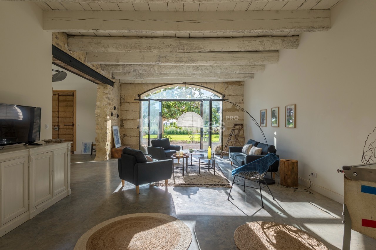 Salon lumineux avec poutres apparentes, mobilier moderne, grande baie vitrée donnant sur un jardin verdoyant.