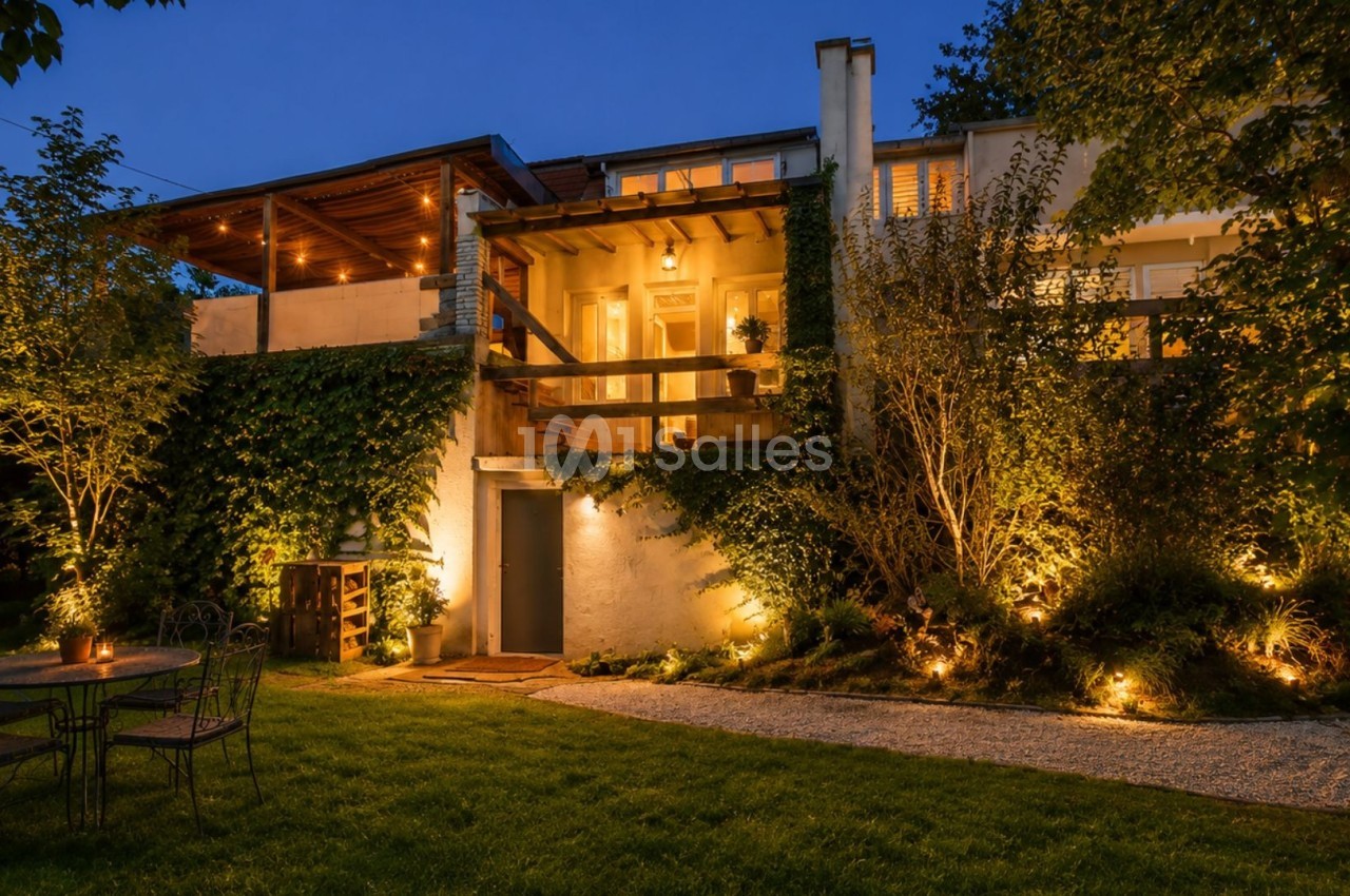 Maison illuminée en soirée avec terrasse en bois, jardin aménagé et mobilier extérieur sur pelouse.