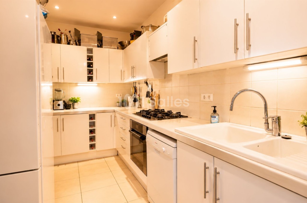 Cuisine moderne avec meubles blancs, évier en inox, plaques de cuisson, réfrigérateur et éclairage chaleureux.
