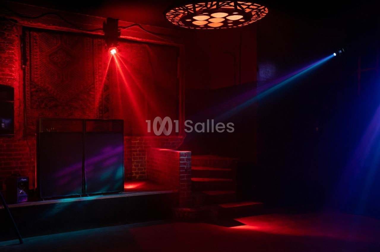 Salle sombre avec éclairage rouge et bleu, une estrade avec équipement audio et un tapis mural en arrière-plan.