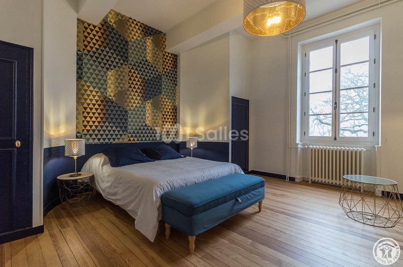 Chambre lumineuse avec un lit double, un banc bleu, un mur décoratif géométrique et une grande fenêtre donnant sur l…