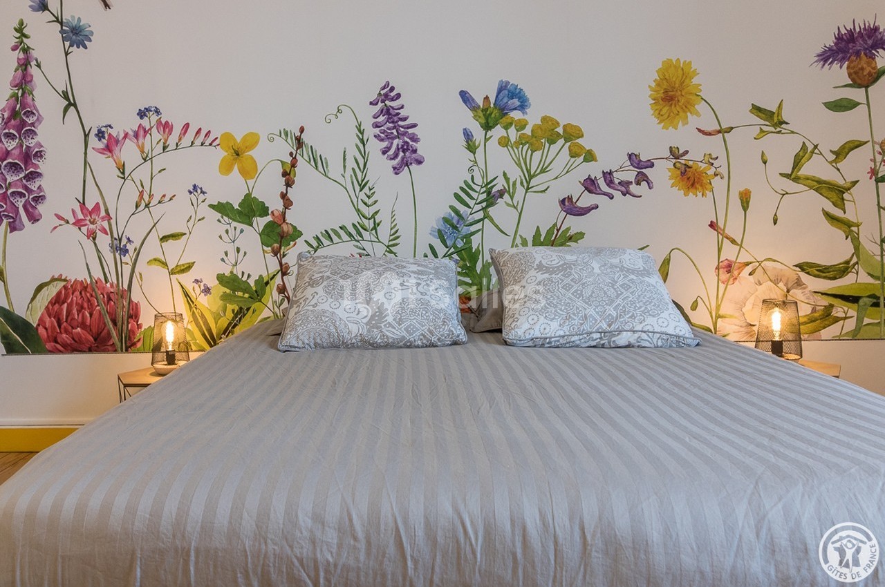 Lit double avec housse grise, encadré de tables de chevet et éclairé par des lampes, devant un mur décoré de fleurs colorées.