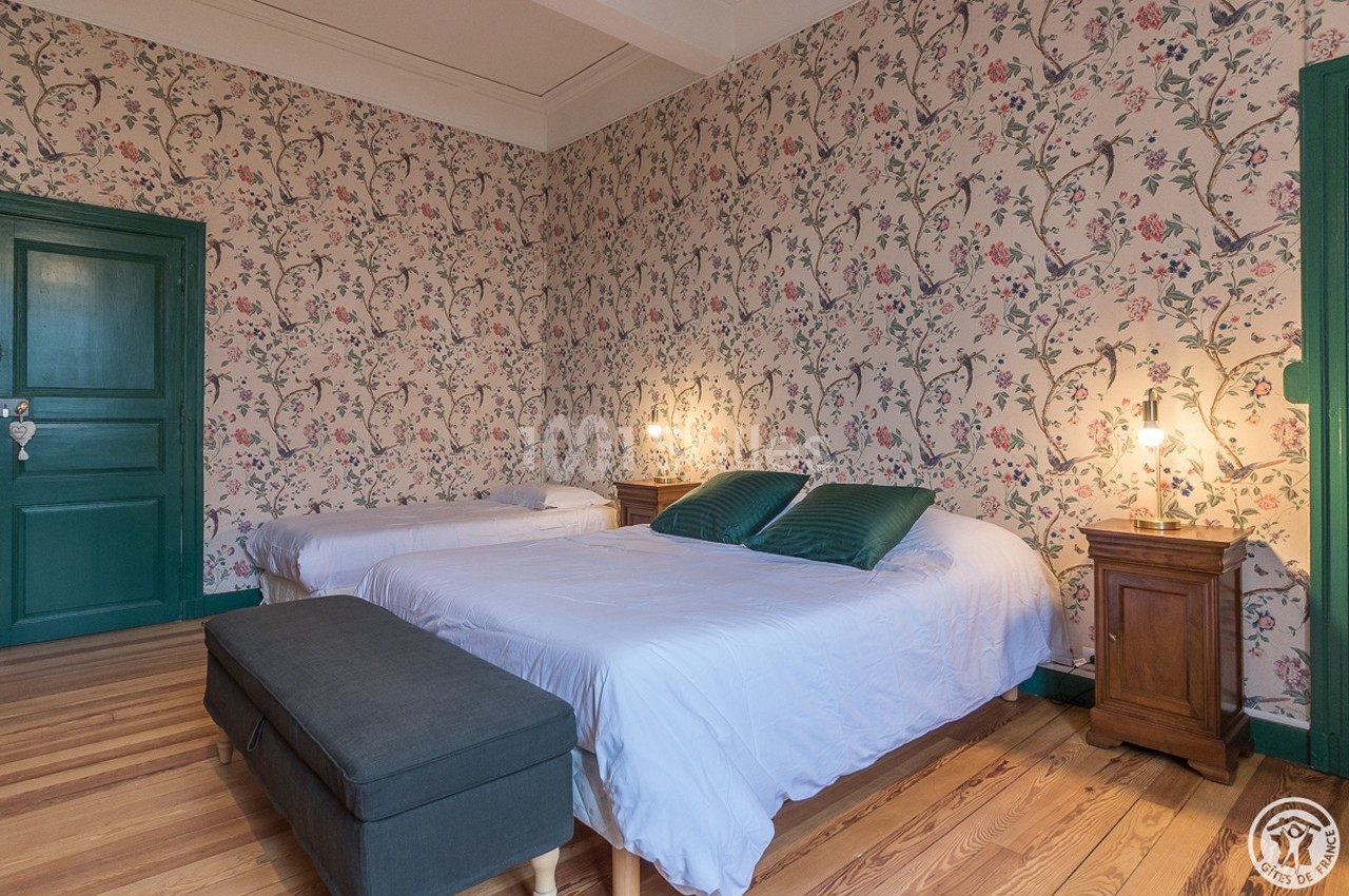 Chambre avec deux lits, parquet en bois, murs tapissés de motifs floraux et mobilier en bois avec lampes de chevet.