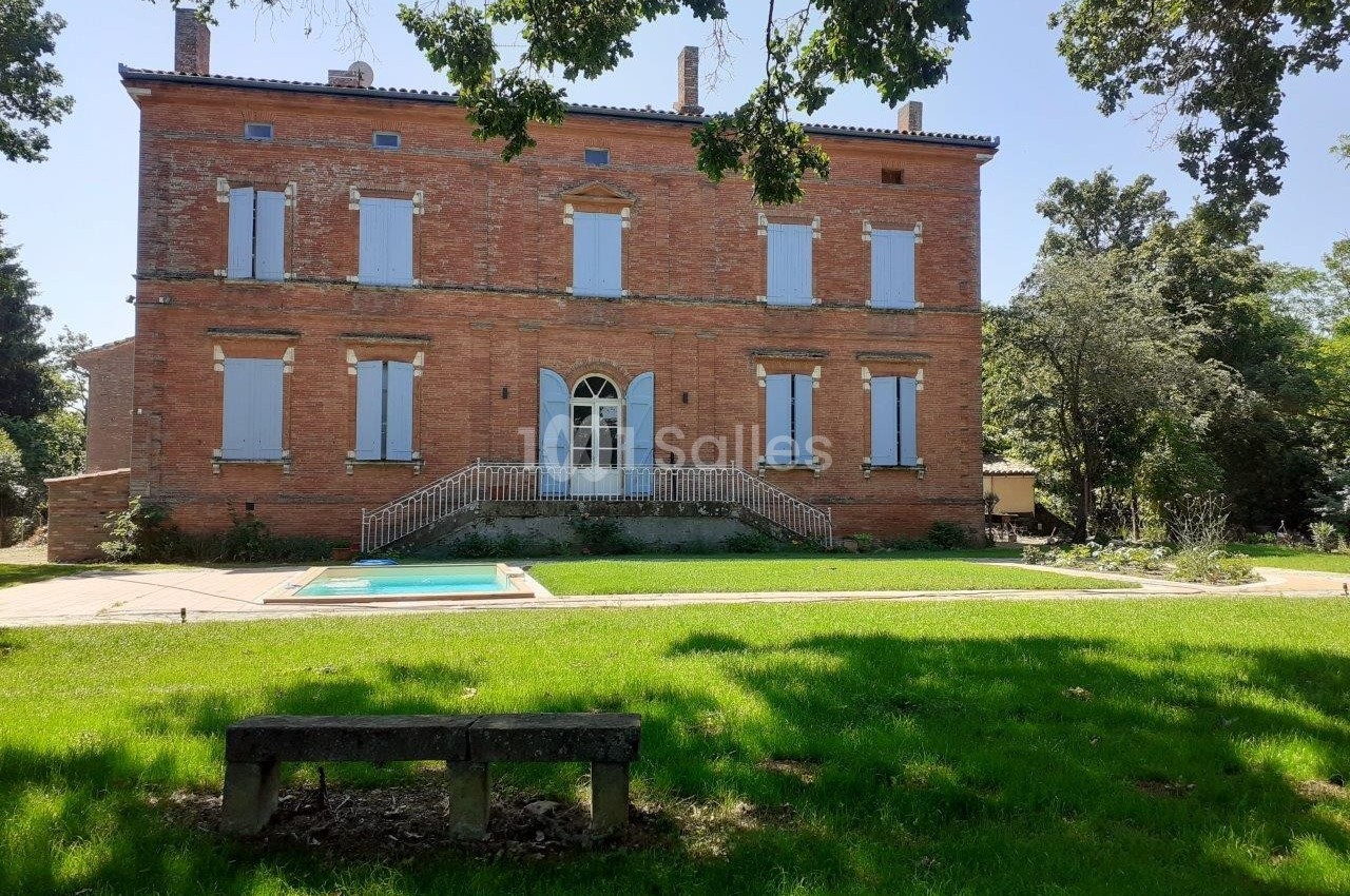 Façade d'une grande maison en briques rouges avec volets fermés, jardin verdoyant, piscine et banc au premier plan.