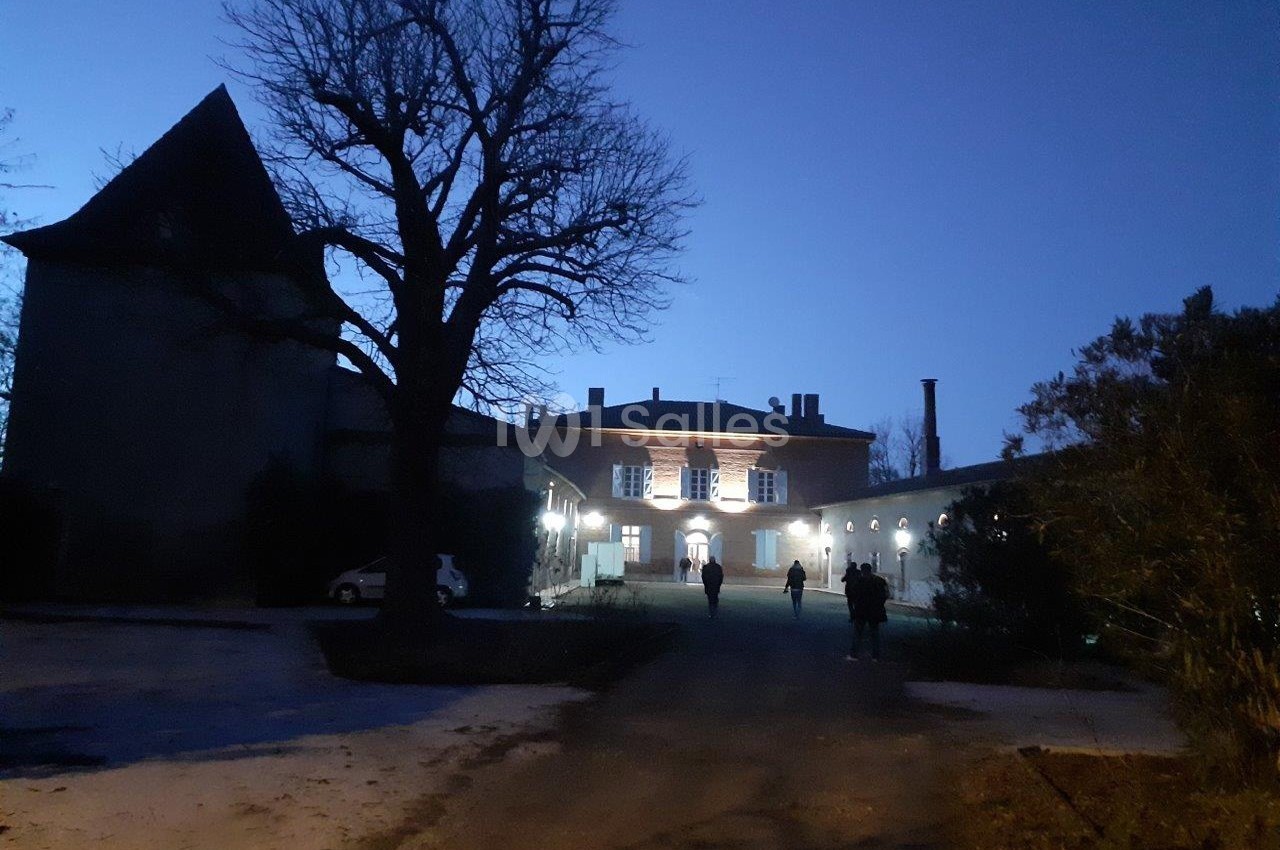 Allée sombre menant à un bâtiment éclairé entouré d'arbres et de silhouettes de personnes au crépuscule.