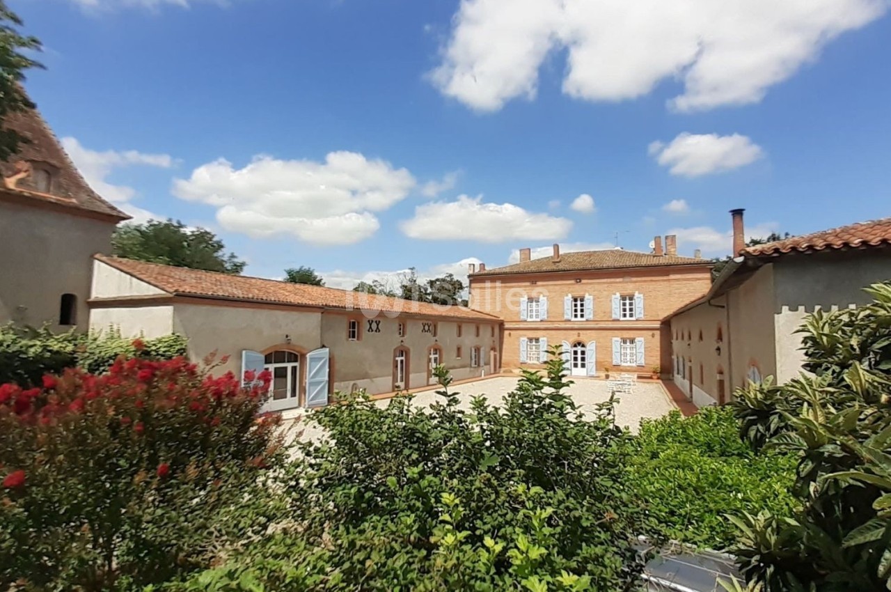 Cour intérieure d'un domaine avec bâtiments en pierre, végétation au premier plan et ciel bleu parsemé de nuages.