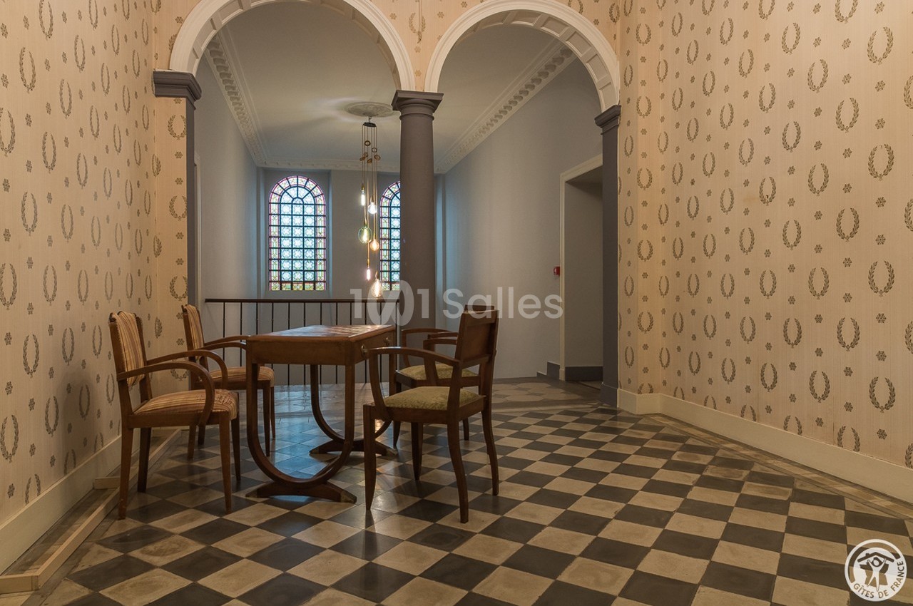 Salle à manger avec table en bois, chaises assorties, carrelage en damier et fenêtres à vitraux colorés.