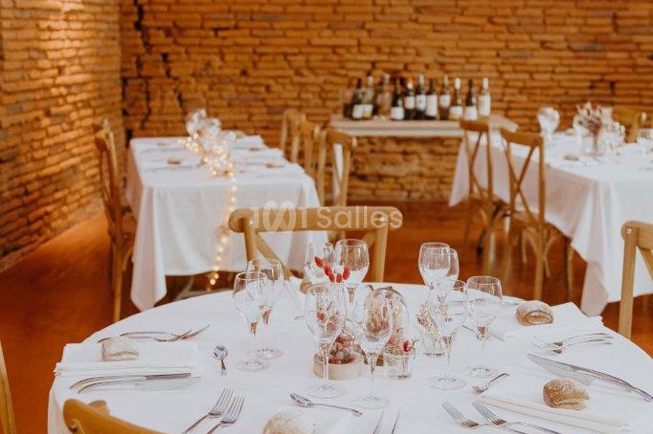 Salle de réception avec tables dressées, nappes blanches, vaisselle élégante et mur de briques en arrière-plan.