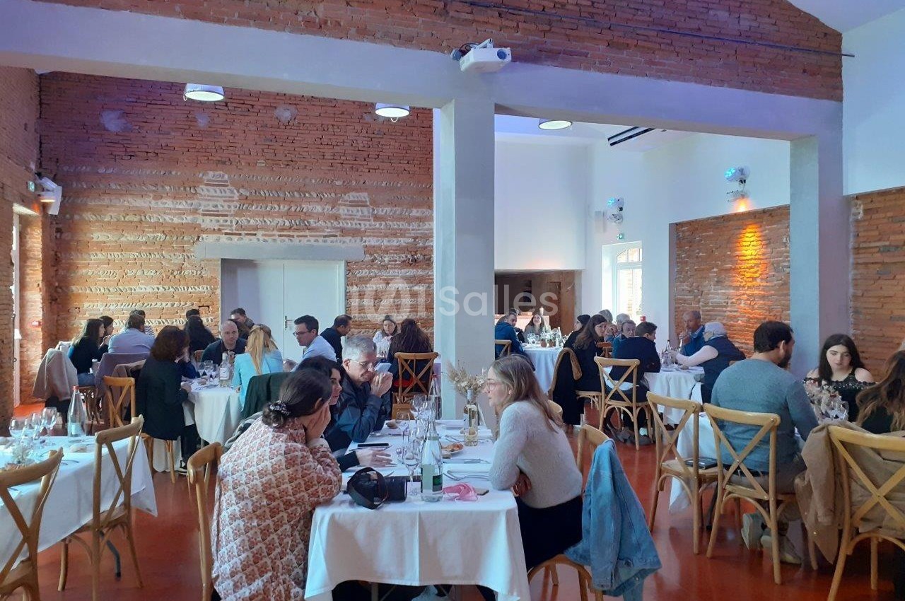Salle de restaurant avec des murs en briques, des tables dressées et des convives en pleine discussion.