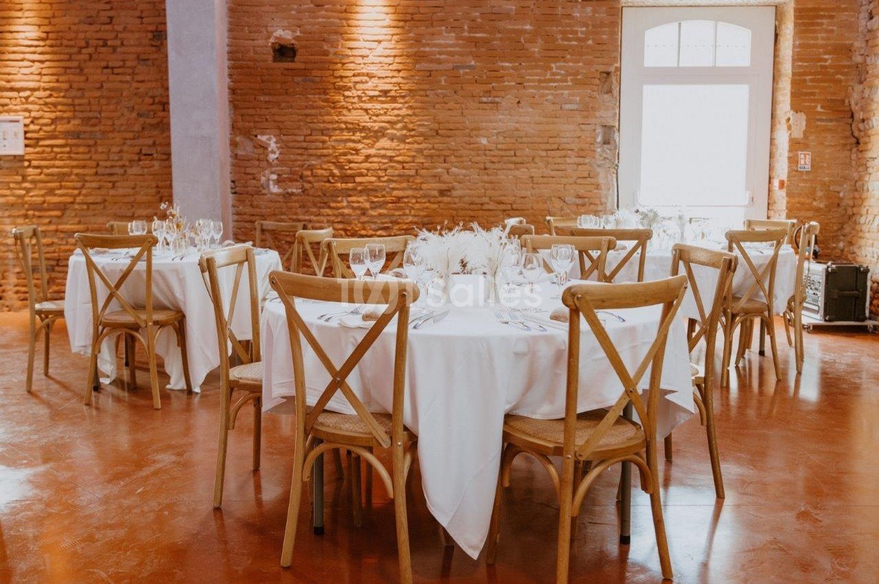 Salle avec des tables rondes dressées, nappes blanches, chaises en bois et murs en briques apparentes.
