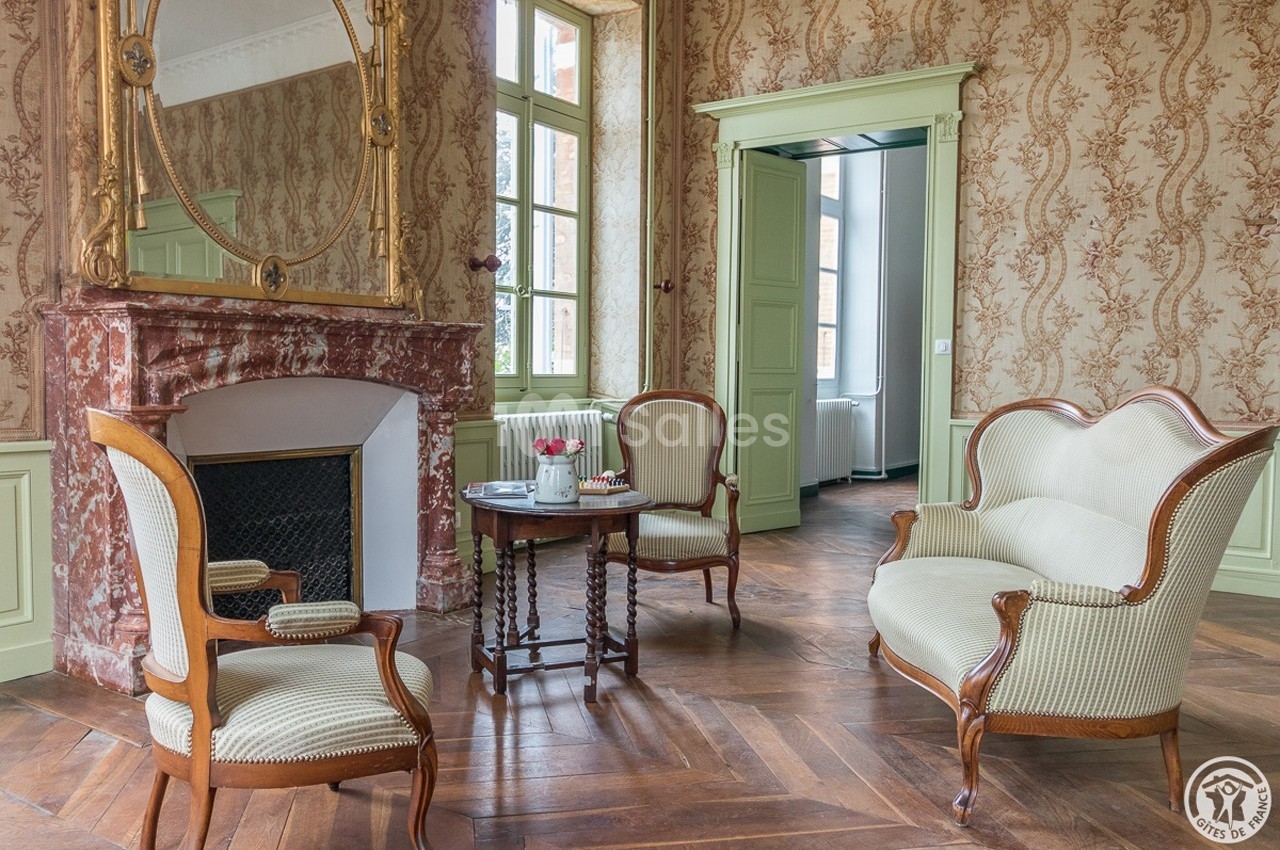 Salon élégant avec cheminée en marbre, meubles anciens en bois et tissu clair, et parquet en chevrons.