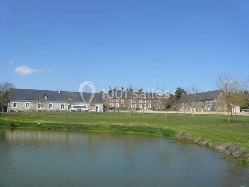 Bâtiments en pierre avec toits en ardoise entourés d'arbres, près d'un étang sous un ciel bleu.