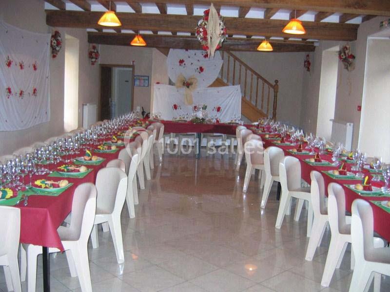 Salle décorée avec des tables alignées, nappes rouges, chaises blanches et vaisselle disposée pour un repas festif.