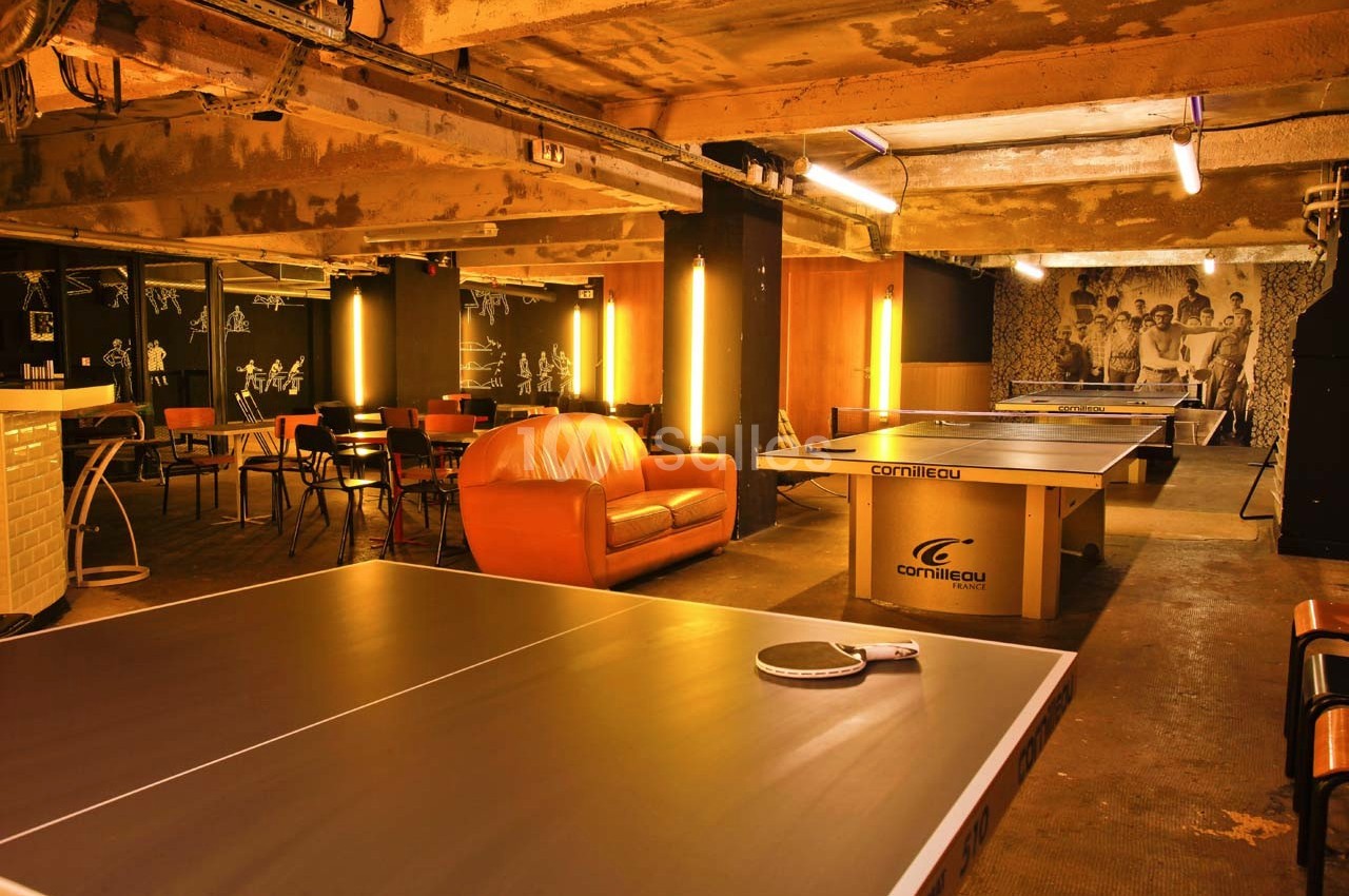 Salle de loisirs avec tables de ping-pong, canapé orange, chaises et éclairage chaleureux dans un espace industriel.