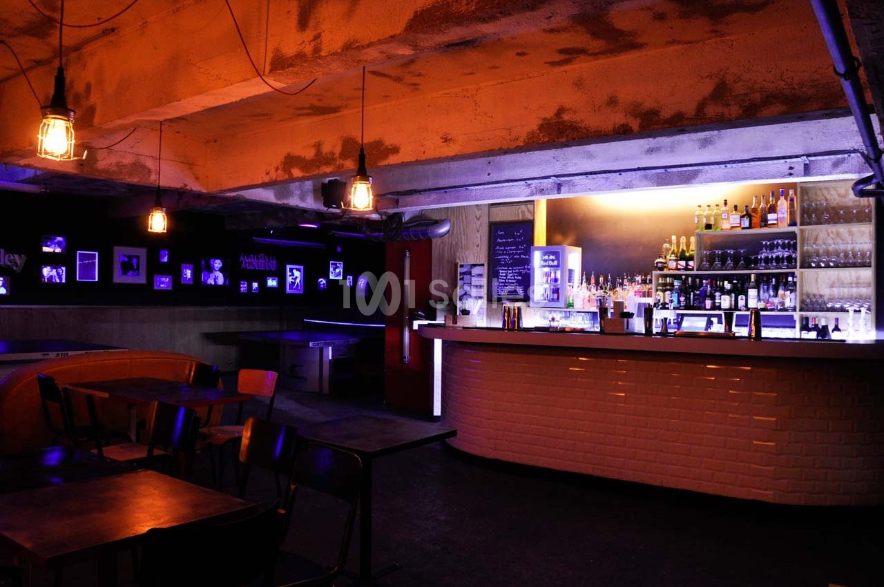 Bar avec éclairage tamisé, comptoir illuminé, étagères de bouteilles et tables vides dans une ambiance industrielle.