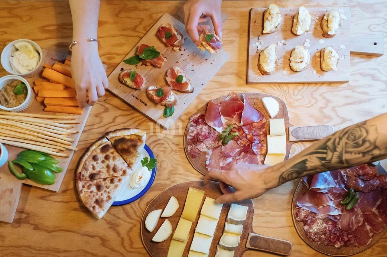 Table en bois garnie de charcuteries, fromages, légumes, pains et pizzas, avec des mains servant les plats.