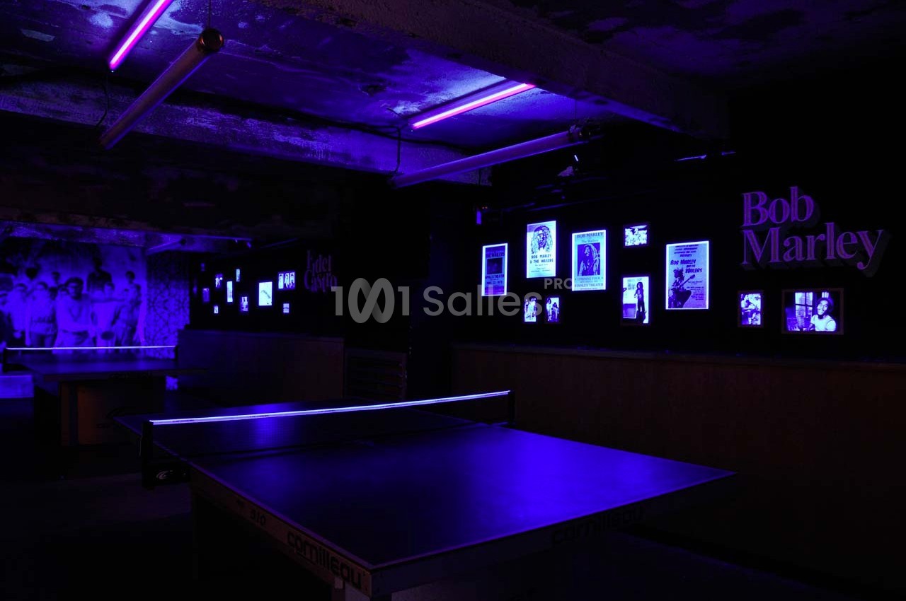 Salle sombre éclairée par des lumières violettes, avec des tables de ping-pong et des affiches de Bob Marley au mur.