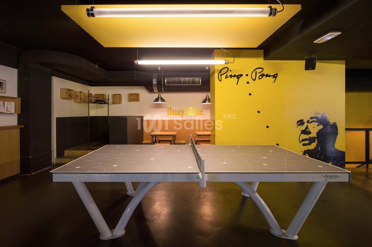 Table de ping-pong grise dans un espace intérieur moderne avec des murs jaunes décorés et un éclairage suspendu.