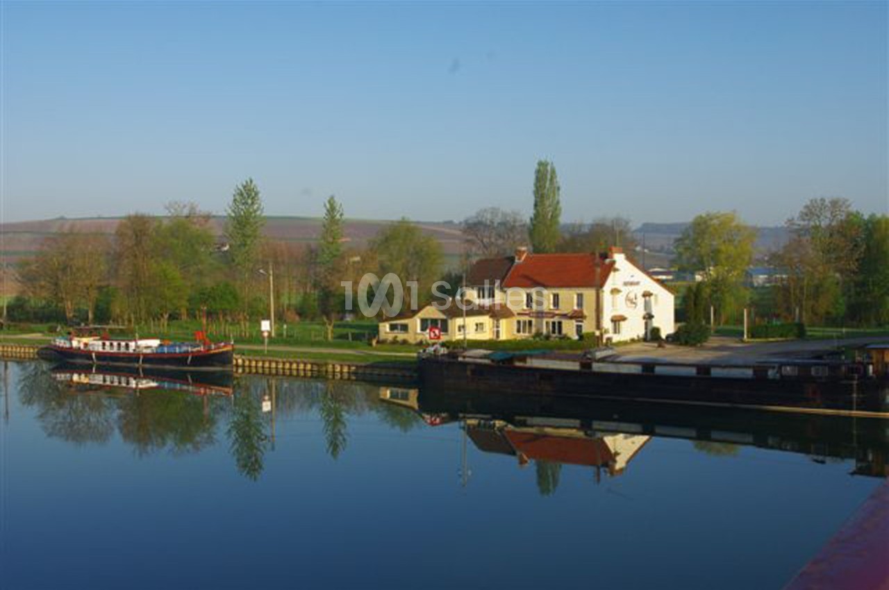 Maison au bord d'un canal calme avec des péniches amarrées, entourée de verdure et d'un paysage rural.