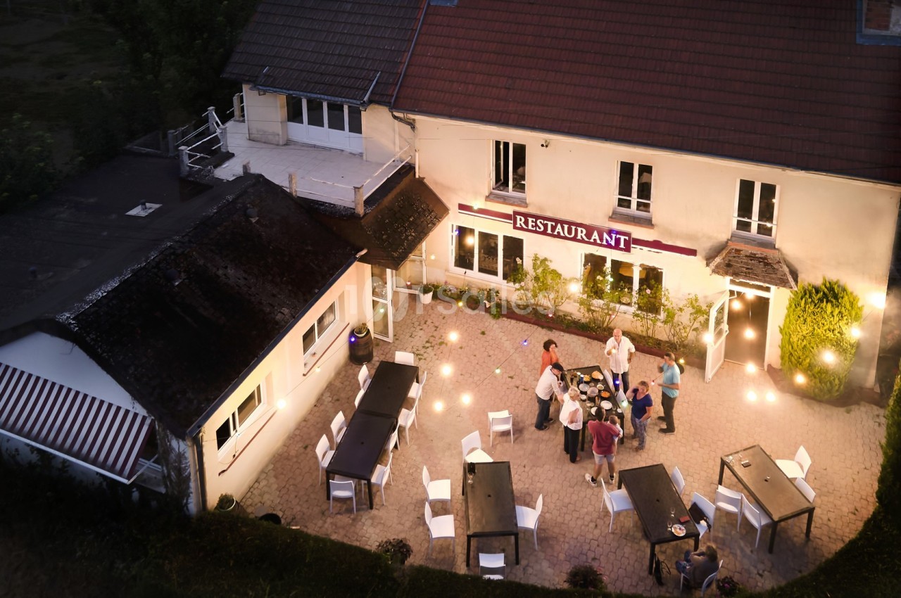 Vue aérienne d'une terrasse de restaurant éclairée en soirée, avec des tables et un groupe de personnes discutant.