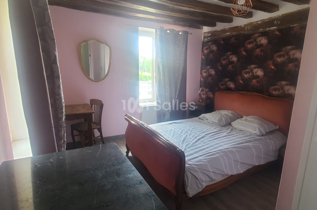 Chambre avec lit double, murs roses, poutres apparentes, table en bois, miroir ovale et fenêtre donnant sur l'extérieur.