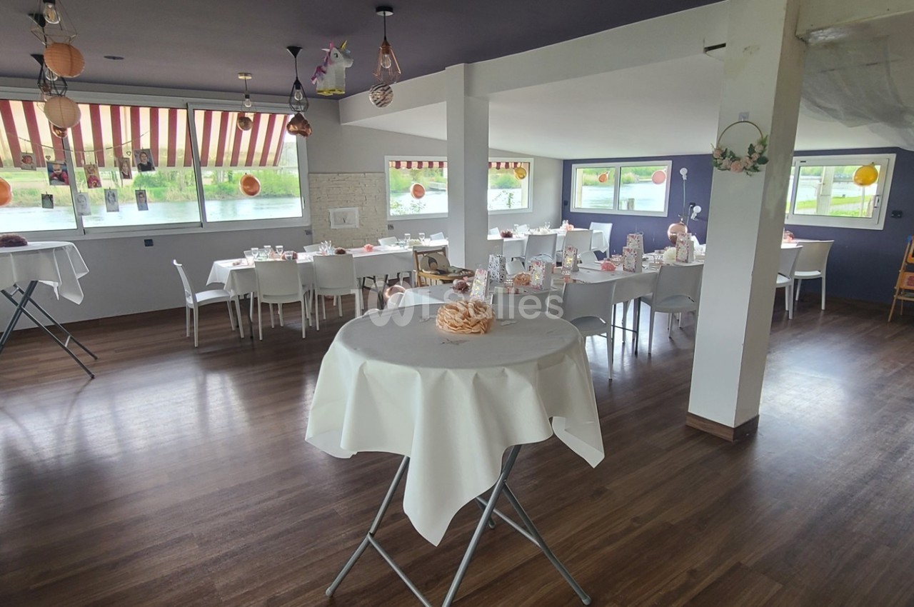 Salle lumineuse aménagée pour un événement, avec tables rondes dressées et décorations suspendues.