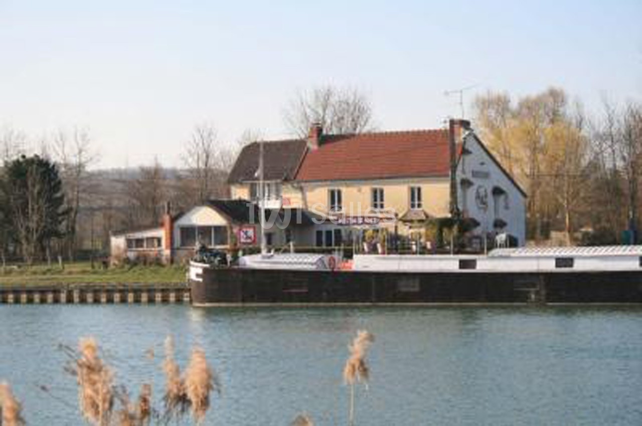 Bâtiment avec terrasse au bord d'un canal, un bateau amarré au premier plan et des arbres en arrière-plan.