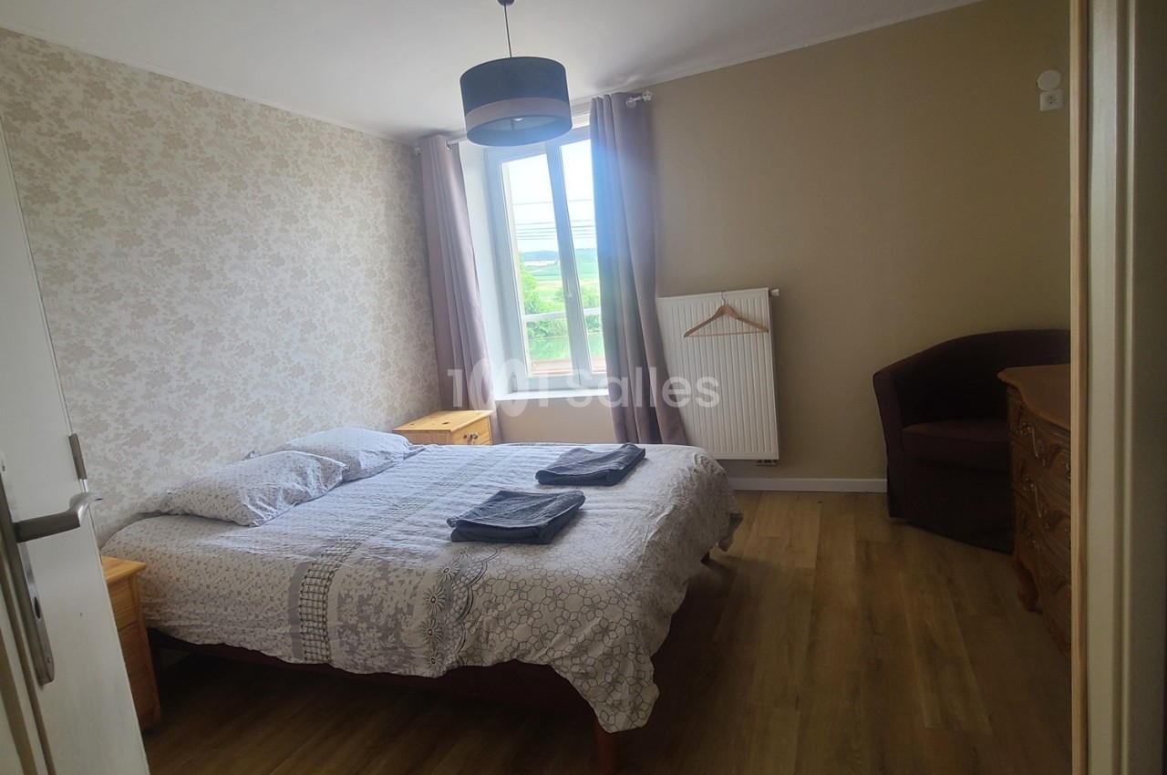 Chambre lumineuse avec lit double, tables de chevet, fauteuil, commode, fenêtre donnant sur un paysage verdoyant.