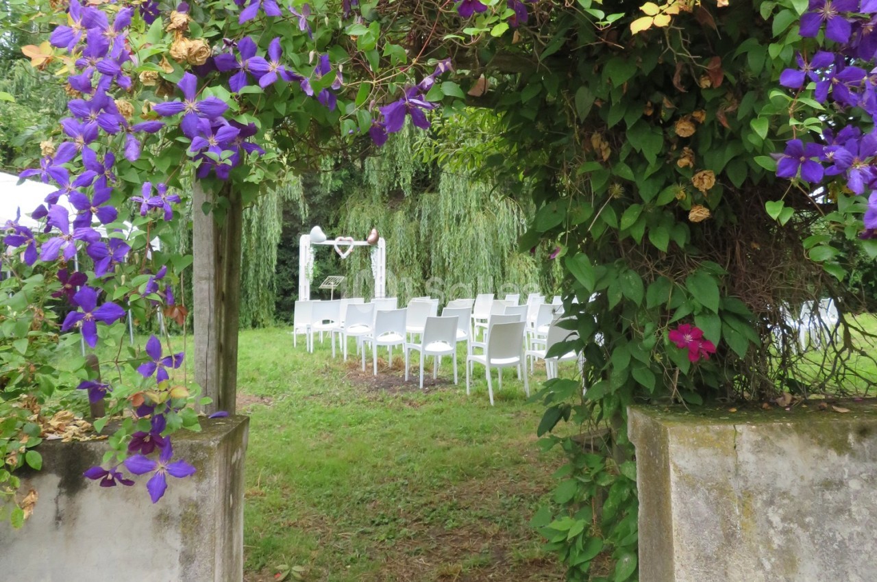 Allée fleurie menant à une cérémonie en plein air avec des chaises blanches et un autel sous un saule pleureur.