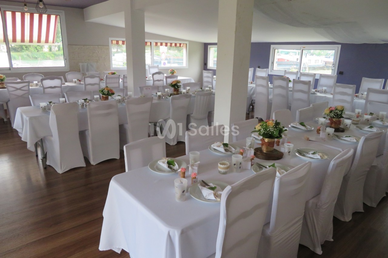 Salle de réception aménagée avec des tables dressées, nappes blanches et décorations florales.
