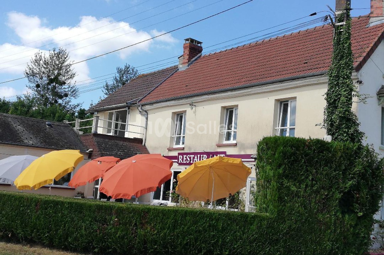 Façade d'un restaurant avec des parasols colorés en terrasse, entourée de haies et sous un ciel dégagé.