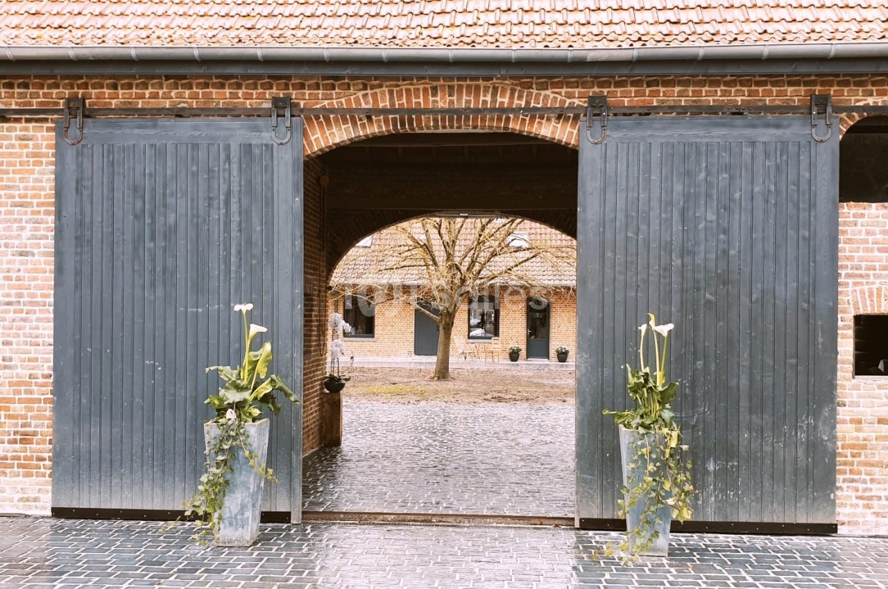 Portail en bois gris ouvert sur une cour pavée, entourée de bâtiments en briques avec des plantes décoratives.
