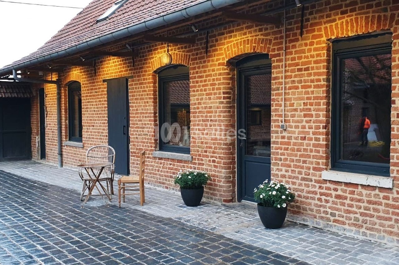 Façade en briques rouges avec fenêtres noires, deux chaises en bois, pots de fleurs et pavés au sol.