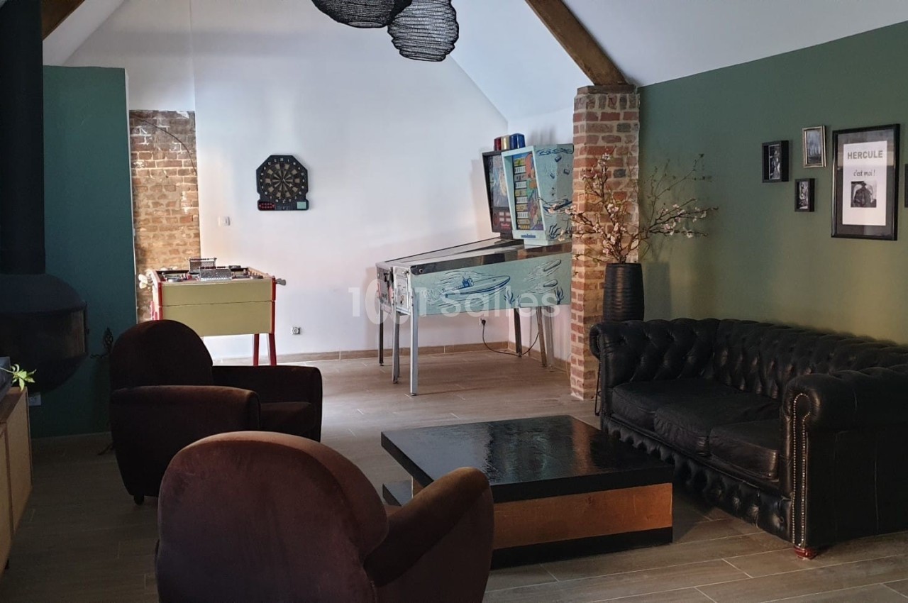 Salon aménagé avec canapé en cuir, fauteuils, poêle, baby-foot, flipper et murs verts décorés de cadres.